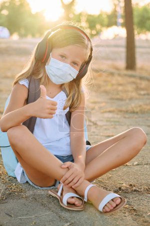Photo for Full body of positive child in protective mask listening to music in headphones and looking at camera while sitting on ground in park during COVID pandemic - Royalty Free Image