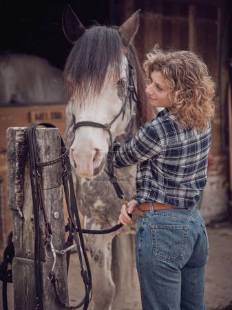 Foto de Vista lateral del contenido propietario femenino de pie cerca de caballo manchado con halter negro en establo en el día de verano en el campo - Imagen libre de derechos
