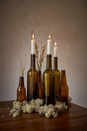 Téléchargez les photos : Ensemble de bouteilles en verre vides avec des bougies blanches placées sur la table avec des fleurs séchées près du mur dans la pièce lumineuse - en image libre de droit