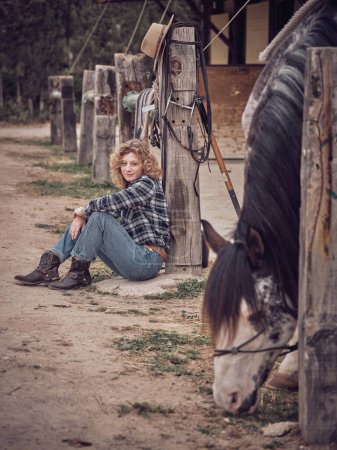 Foto de Cuerpo completo de encantadora hembra sentada cerca de un pilar de madera mientras mira a un lindo caballo con brida en el establo en el día de verano - Imagen libre de derechos