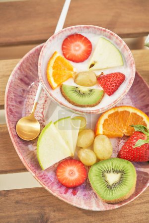 Foto de De arriba cuenco de yogur cubierto con una variedad de frutas y bayas servidas en bandeja de cerámica con cuchara durante el desayuno en la cocina - Imagen libre de derechos