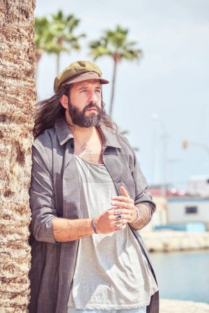 Téléchargez les photos : Homme adulte aux cheveux longs en tenue élégante appuyé sur un palmier sur un rivage sablonneux près d'un quai - en image libre de droit