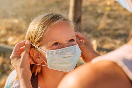 Photo for Crop unrecognizable person putting on protective mask on face of positive preteen child while standing on pathway in woods during COVID 19 pandemic - Royalty Free Image