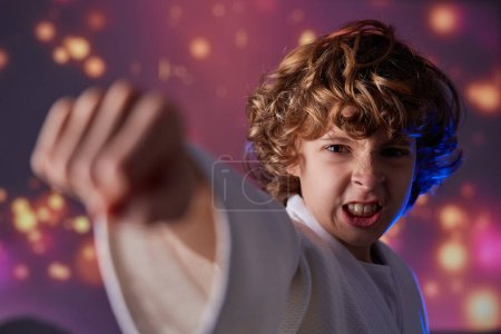 Téléchargez les photos : Garçon martial aux cheveux bouclés en kimono blanc pratiquant des coups de poing lors d'un entraînement intense de judo en studio avec des lumières brillantes - en image libre de droit