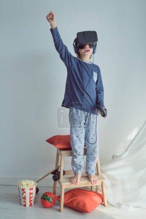 Téléchargez les photos : Corps complet d'un enfant anonyme pieds nus en pyjama levant le bras avec la bouche ouverte tout en jouant à un jeu de réalité virtuelle portant un casque et debout sur une échelle contre un mur blanc - en image libre de droit