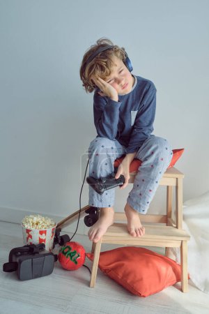 Photo for Full body of little blond haired barefoot unhappy boy in pajama and headphones sitting with head in hand while playing video game with joystick and losing against white background - Royalty Free Image