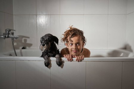 Foto de Niño contento mirando hacia otro lado con mirada difícil mientras está sentado en una bañera blanca con perro durante la rutina de higiene en el baño ligero - Imagen libre de derechos