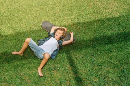 Photo for From above of excited preteen child shouting and listening to music in headphones while lying on skateboard on grassy lawn - Royalty Free Image