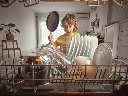Foto de Niño contento con sartén en las manos de pie cerca de lavavajillas abierto con vario vajilla limpia durante la rutina del hogar en la cocina - Imagen libre de derechos