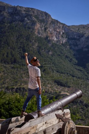 Foto de Vista posterior cuerpo completo de turista masculino en traje casual y sombrero de pie en pistola vieja con el brazo levantado mientras disfruta de pintoresco paisaje montañoso - Imagen libre de derechos