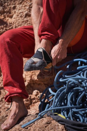 Téléchargez les photos : Crop anonyme pieds nus alpiniste mettre des chaussures d'escalade tout en étant assis sur un sol rocheux rugueux près de cordes dans la nature le jour de l'été - en image libre de droit