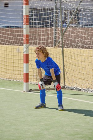 Photo for Full body of preteen goalkeeper in activewear leaning on knees and looking forward while playing soccer during training on sports ground - Royalty Free Image