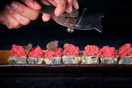 Recadrer un chef anonyme tranchant de la truffe noire sur des rouleaux de sushi tempura avec du thon servi sur assiette sur fond noir au restaurant