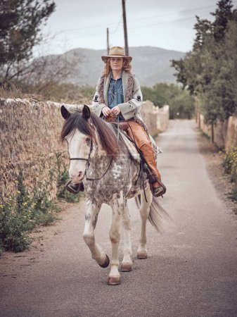 Téléchargez les photos : Femme en chapeau équitation mignon cheval sur route asphaltée le long du mur de pierre dans la zone rurale de la campagne le jour de l'été - en image libre de droit