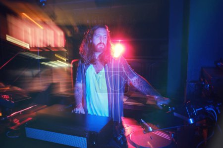 Photo for Talented male in casual wear standing at professional DJ setup while playing music in dark nightclub with long exposure lights - Royalty Free Image
