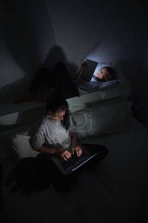 Foto de Alto ángulo de la madre sentada en la cama y el ordenador portátil de navegación, mientras que el niño viendo vídeo en la tableta y sonriendo en el dormitorio por la noche - Imagen libre de derechos