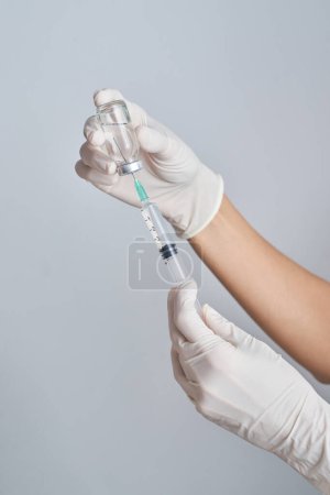 Téléchargez les photos : Mains de culture médecin méconnaissable dans des gants de protection établissant le vaccin du flacon à la seringue sur fond blanc dans la pièce - en image libre de droit
