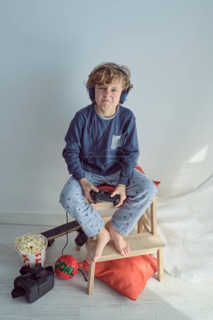 Photo for Full length from above of disappointed blond haired kid in home wear and headphones losing in video game while playing with joystick sitting on stool and looking at camera - Royalty Free Image