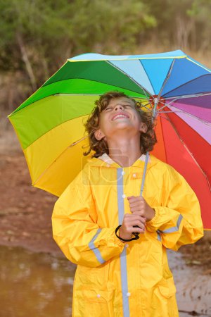 Foto de Niño alegre con paraguas de arco iris vistiendo impermeable amarillo de pie con los ojos cerrados en charco en el bosque - Imagen libre de derechos