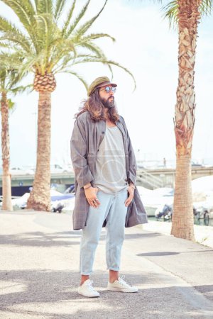 Foto de Confiado hipster en gorra y gafas de sol caminando en puerto con palma cerca del río con embarcaciones amarradas en día soleado en la ciudad - Imagen libre de derechos