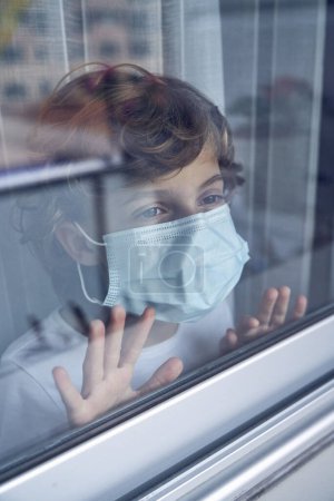Photo for Through glass view of kid wearing protective face mask standing with hands on window and looking away thoughtfully while staying at home during COVID 19 pandemic - Royalty Free Image