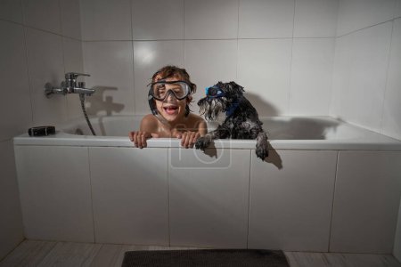 Foto de Niño positivo en máscara de buceo sentado cerca de lindo perro negro en bañera blanca mientras juega en el baño ligero en casa - Imagen libre de derechos