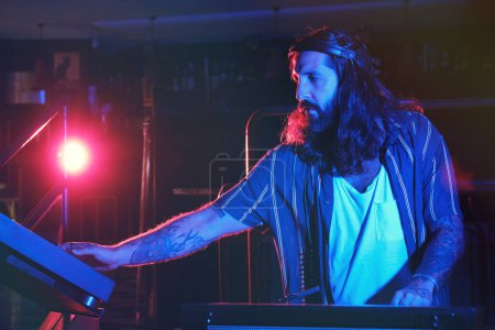 Téléchargez les photos : DJ masculin talentueux dans les écouteurs jouant de la musique sur mélangeur audio moderne et CDJ tout en travaillant dans une boîte de nuit avec un éclairage tamisé - en image libre de droit