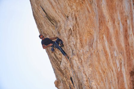 Photo for From below full body of male alpinist in safety harness ascending on rough rocky cliff during training on summer day - Royalty Free Image