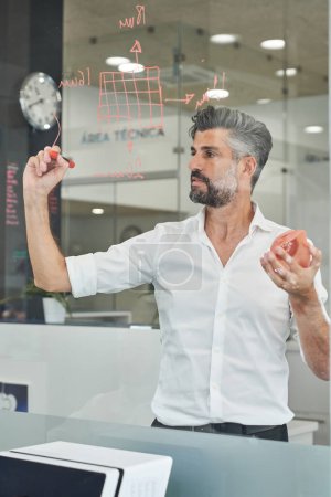 Foto de Ventana de canal de ingeniero masculino serio en desgaste formal tomando notas en la pared de vidrio al diseñar el modelo de máscara facial - Imagen libre de derechos
