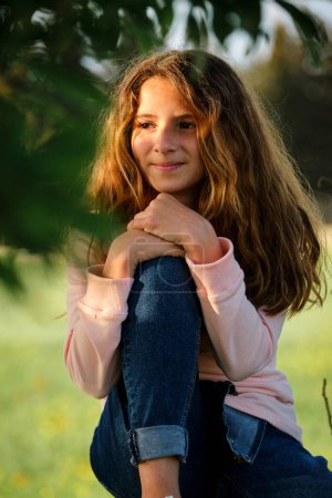 Photo for Girl leaning on knee and looking away while relaxing under lush tree branches on sunny day on summer day - Royalty Free Image
