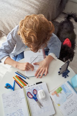 Photo for From above of anonymous boy sitting at desk together with dog while erasing notes in notepad during lesson via smartphone at home - Royalty Free Image