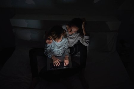 Photo for From above of woman hugging son typing on laptop while doing homework together on bed in dark bedroom at home - Royalty Free Image
