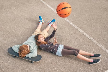 Foto de Desde arriba feliz adolescente lanzando pelota de baloncesto mientras se encuentra en el campo de deportes cerca de amigo con monopatín - Imagen libre de derechos