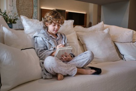 Photo for Full body of curly haired boy with barefoot crossed legs sitting on sofa with cushions and drawing sketch in paper notebook in living room - Royalty Free Image