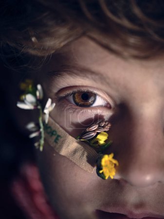 Téléchargez les photos : La moitié du visage d'un garçon mignon avec une fleur sur le visage et un pansement avec un mot d'espoir regardant loin dans la pièce lumineuse - en image libre de droit