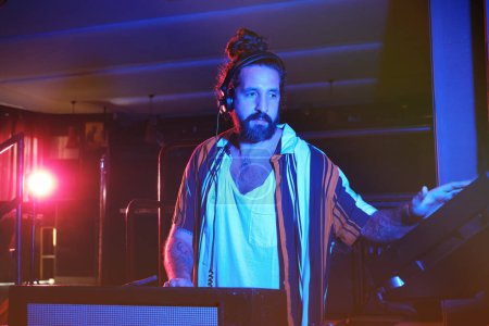 Photo for Talented bearded male DJ in headphones playing music on modern professional equipment while performing song in nightclub with dim light - Royalty Free Image