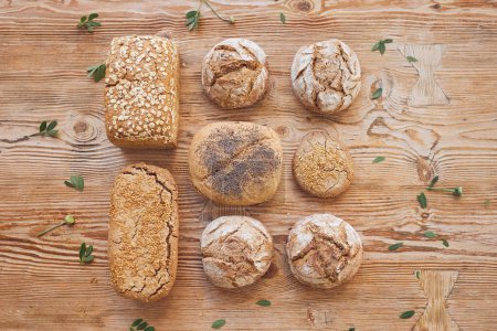 Foto de Colocación plana de apetitosos panes crujientes panes y panecillos colocados sobre una mesa de madera en hileras y decorados con hojas verdes en panadería - Imagen libre de derechos