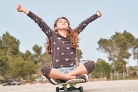 Téléchargez les photos : Niveau du sol du patineur adolescent heureux assis sur le skateboard avec les bras tendus et jouissant de la liberté - en image libre de droit