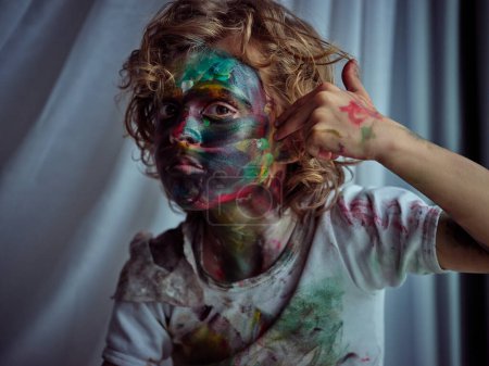 Foto de Rebelde chico sucio con el pelo rizado untando pintura colorida en la cara mientras mira a la cámara con mirada seria en el estudio de luz - Imagen libre de derechos