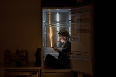 Foto de Vista lateral del chico serio en ropa casual sentado en nevera con las piernas cruzadas y el uso de la tableta en la cocina oscura en casa - Imagen libre de derechos