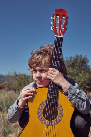Téléchargez les photos : Talentueux petit garçon guitariste aux cheveux blonds bouclés dans des vêtements décontractés embrassant l'instrument et regardant loin rêveur sous un ciel bleu sans nuages à la campagne - en image libre de droit