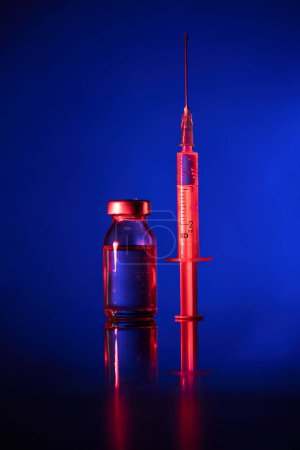 Foto de Ampolla médica y jeringa rellenadas con la vacuna COVID 19 colocadas sobre superficie reflectante en iluminación de neón sobre fondo azul - Imagen libre de derechos