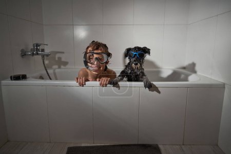 Foto de Niño positivo en máscara de buceo con perro en gafas de sol sentado en la bañera mientras juega en el baño durante la rutina de higiene diaria - Imagen libre de derechos