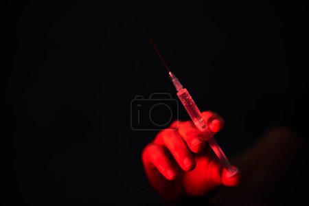 Photo for Unrecognizable person holding medical syringe with needle filled with dose of COVID 19 vaccine in red lighting - Royalty Free Image