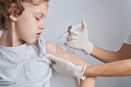Foto de Recorte médico irreconocible en guantes de látex inyectando la vacuna en el hombro del niño sobre fondo blanco durante la epidemia en la sala de luz - Imagen libre de derechos