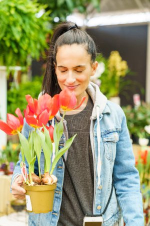 Téléchargez les photos : Femme hispanique positive aux cheveux noirs et aux yeux fermés tenant un pot avec des tulipes rouges en fleurs tout en se tenant debout dans un magasin floral léger - en image libre de droit