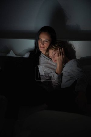 Photo for Caring woman stroking face of sleeping son while helping to do homework with laptop on bed in bedroom late at night - Royalty Free Image