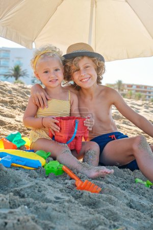 Foto de Niño feliz con sombrero abrazando a la niña en traje de baño amarillo y mirando a la cámara mientras está sentado en la playa de arena - Imagen libre de derechos