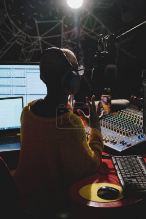 Photo for Back view of anonymous ethnic female radio host in headphones recording podcast while sitting at table in dark broadcast studio - Royalty Free Image
