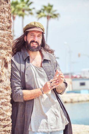 Foto de Alegre hombre barbudo con ropa hipster apoyado en la palmera y mirando a la cámara en la orilla arenosa cerca del muelle - Imagen libre de derechos